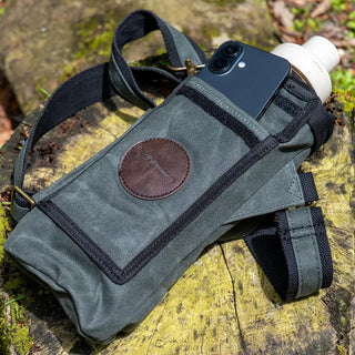 A green waxed canvas water bottle pouch, with extra pockets for other items, shown here with a mobile phone, featuring a shoulder strap.