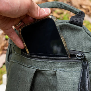 Image shows a mobile phone being placed into a zipped pocket on a green waxed canvas rucksack
