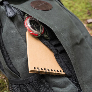 Image shows a notebook and a compass in the front zipped storage pocket of the waxed canvas Quest rucksack