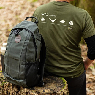 Green waxed canvas rucksack with black mesh side pockets, and a leather logo patch, seen here with a water bottle fitted placed next to a model.
