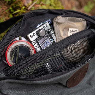 An open waxed canvas sling pouch revealing some suggested contents. A compass, dictaphone, and some money.