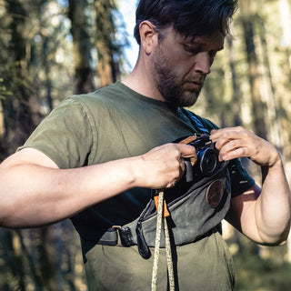 A Green waxed canvas pouch designed to be worn diagonally across the body, modelled with a man removing a camera.