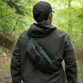 A green waxed canvas sling bag, modelled diagonally across a model's back.