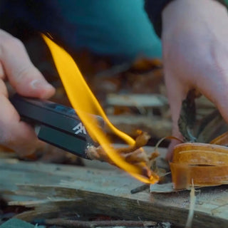 Forest Fundamentals Stealth Firelighter, a ferrocerium rod, in an aluminium holder, with fire lighting cord, striker, and lanyard. The cord is lit and lighting some Birch bark.
