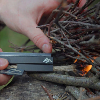 Forest Fundamentals Stealth Firelighter, a ferrocerium rod, in an aluminium holder, with fire lighting cord, striker, and lanyard. The fire cord is lit and is being used to light some Birch twigs.